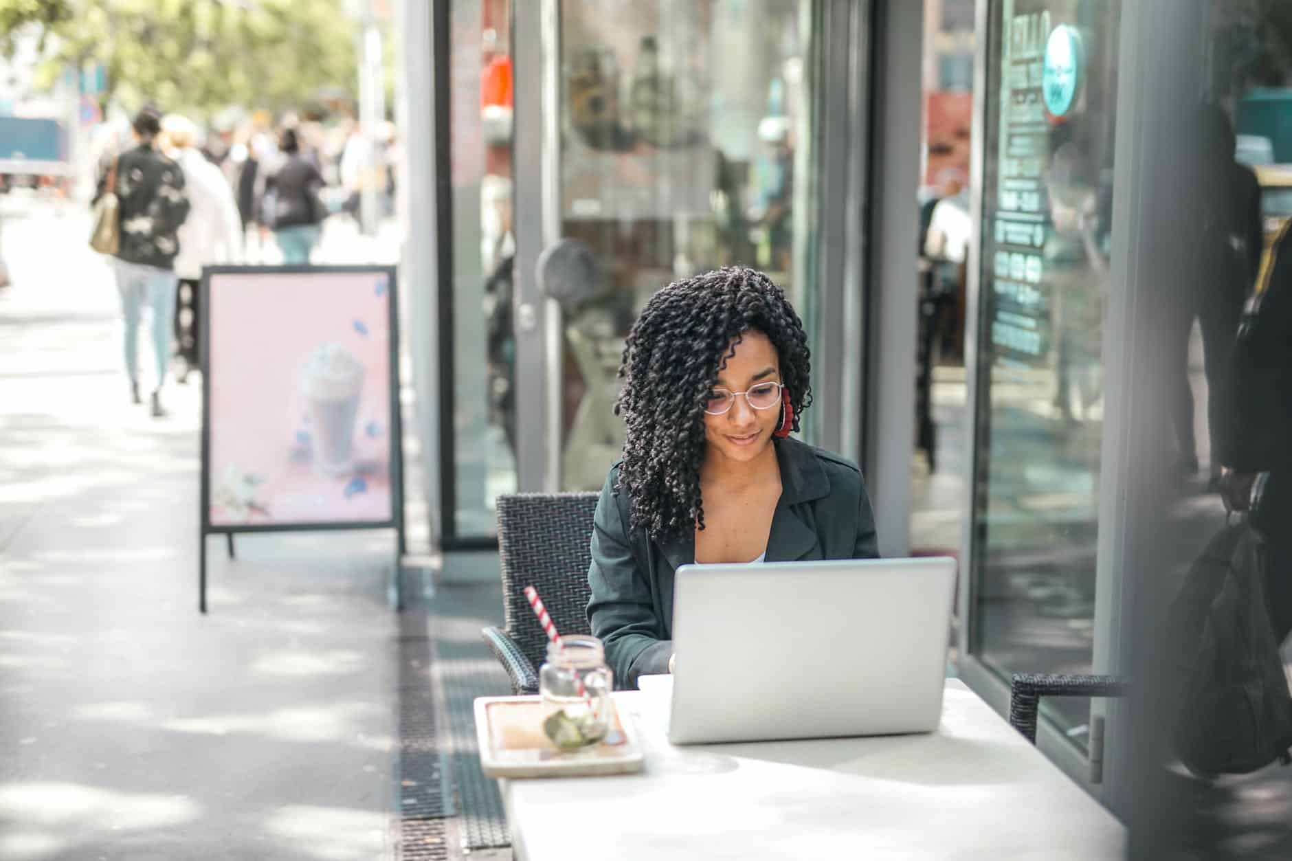 ethnic young woman using laptop while having tasty beverage in modern street cafe working on client management